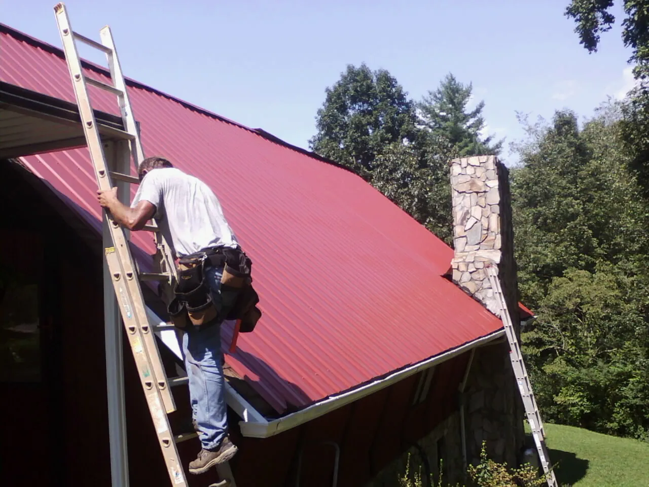 Roofer on a ladder beside a red metal roof and stone chimney — Soffit & Fascia Repair by Roofing Brothers in Montpelier Station